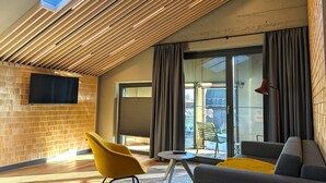 Een moderne woonkamer met een schuine plafond, een flatscreen televisie aan een bakstenen muur, een gele stoel, een grijze bank en een rode vloerlamp.