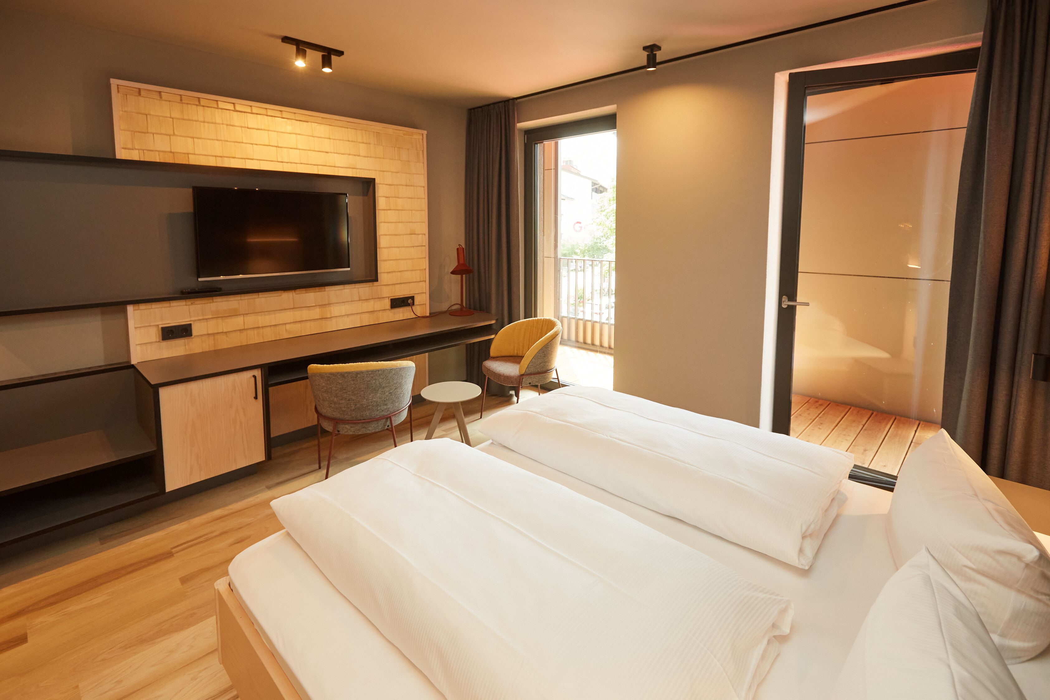 Een moderne hotelkamer met een groot bed, een bureau met stoel, een televisie en een schuifpui naar een balkon.