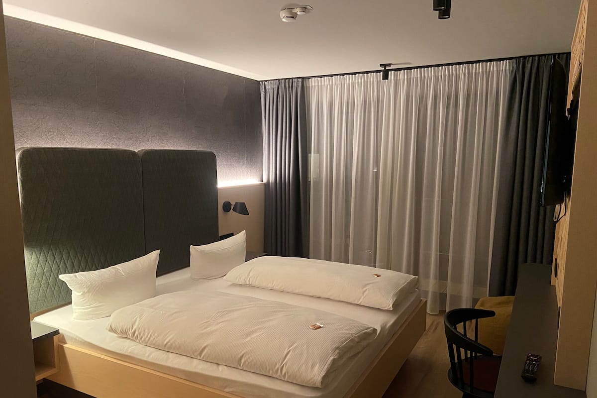 A hotel room with a large bed, a headboard, a bedside table, a lamp, a chair, and a curtain.