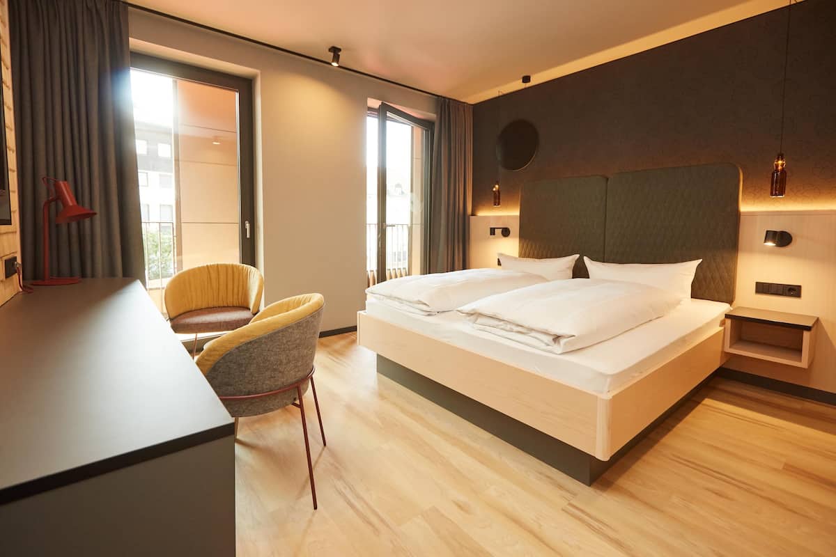 A modern hotel room with a large bed, a desk, a chair, and a view of the city through the window.