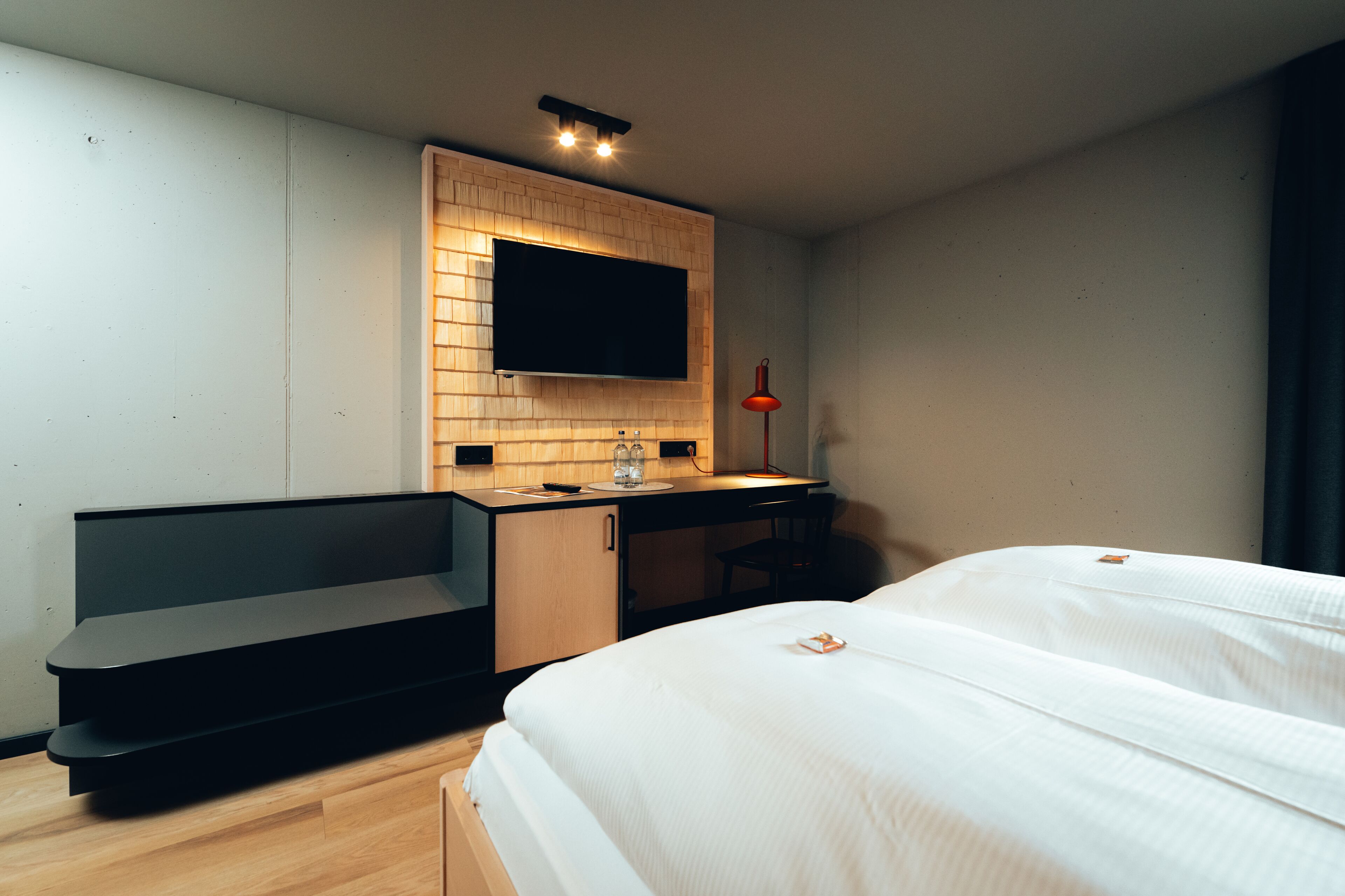 Una habitación de hotel moderna con una cama, un escritorio, un televisor montado en un panel de madera y una lámpara de pie roja.