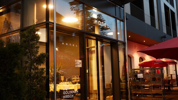 Een modern hotel met een glazen gevel, een zichtbaar restaurant genaamd "Goldenen Hirsch" en terras met parasols.