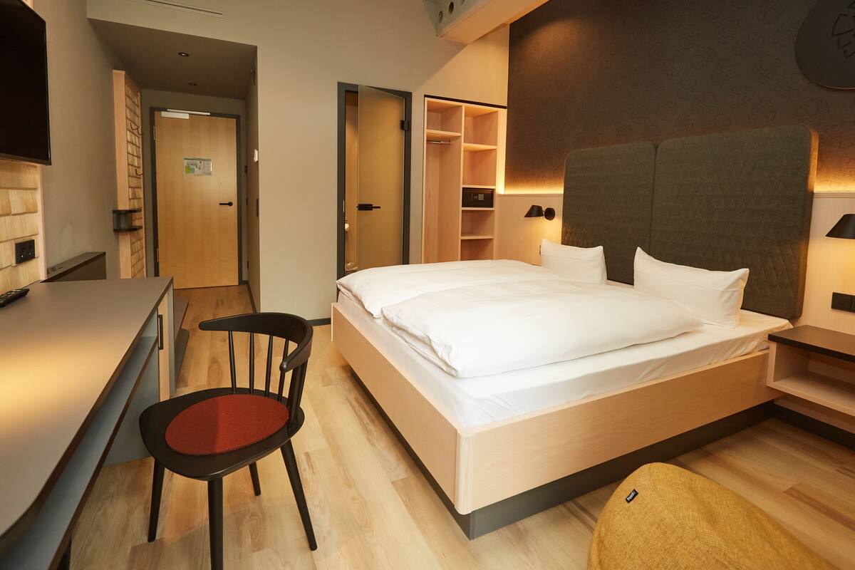 A modern hotel room with a bed, a desk, a chair, and a wardrobe.