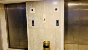 Zona de ascensores con puertas metálicas, un cesto de basura y paneles de control en la pared.