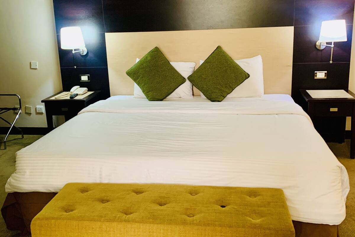 Habitación de hotel con una cama grande, dos almohadas verdes, una mesita de noche con una lámpara y una pequeña mesa con un teléfono.