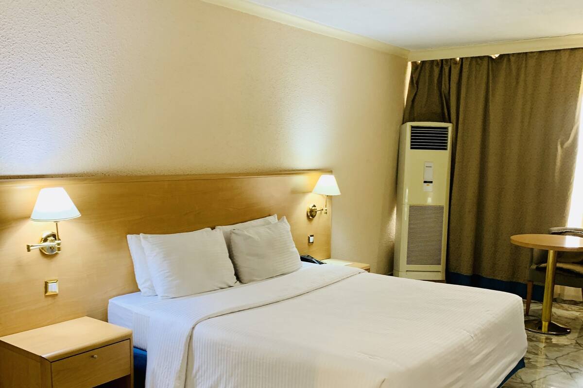 Una habitación de hotel con cama, mesitas de noche, lámparas, un equipo de aire acondicionado y una pequeña mesa.