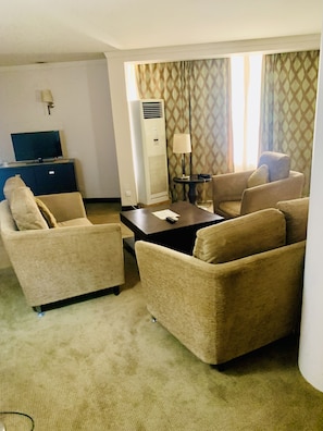 A hotel room with beige sofas, a small table, a TV on a cabinet, and an air conditioner.