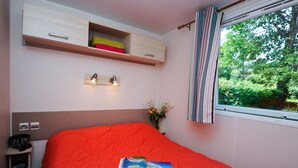 A bedroom with a bed, a bedside table, a shelf with towels, a window with curtains, and a view of greenery outside. - Mobil-home 3 Chambres 31m² (Telgruc-sur-Mer)