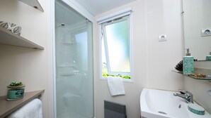 A bathroom with a glass shower enclosure, a window with a towel hanging, a sink with a mirror, and a shelf with decorative items. - Mobil-home 3 Chambres 31m² (Telgruc-sur-Mer)