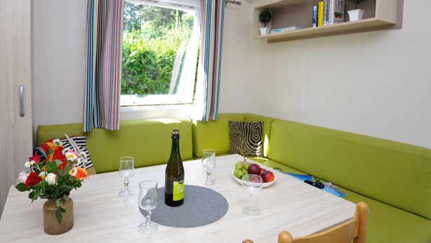 A dining area with a table set for two, a bottle of wine, and a bowl of fruit. - Mobil-home 3 Chambres 31m² (Telgruc-sur-Mer)