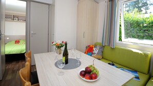 A compact room with a dining table, a green sofa, a window with curtains, and a bed with a green bedspread. - Mobil-home 3 Chambres 31m² (Telgruc-sur-Mer)