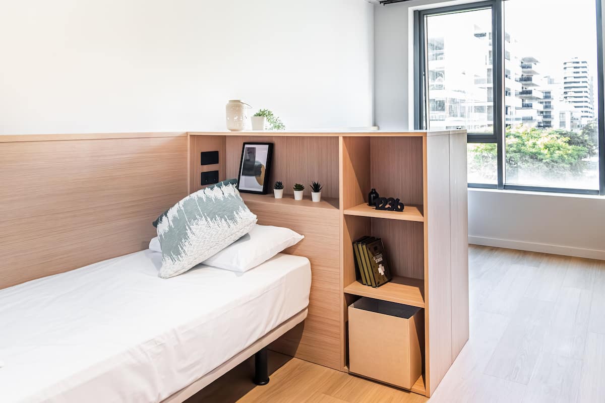 A compact bedroom with a bed, a small shelf with decorative items, and a window with a city view.