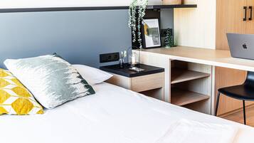 Ein modernes Schlafzimmer mit einem Bett, einem Schreibtisch mit Laptop, einem Nachttisch mit Lampe und einer kleinen Pflanze.