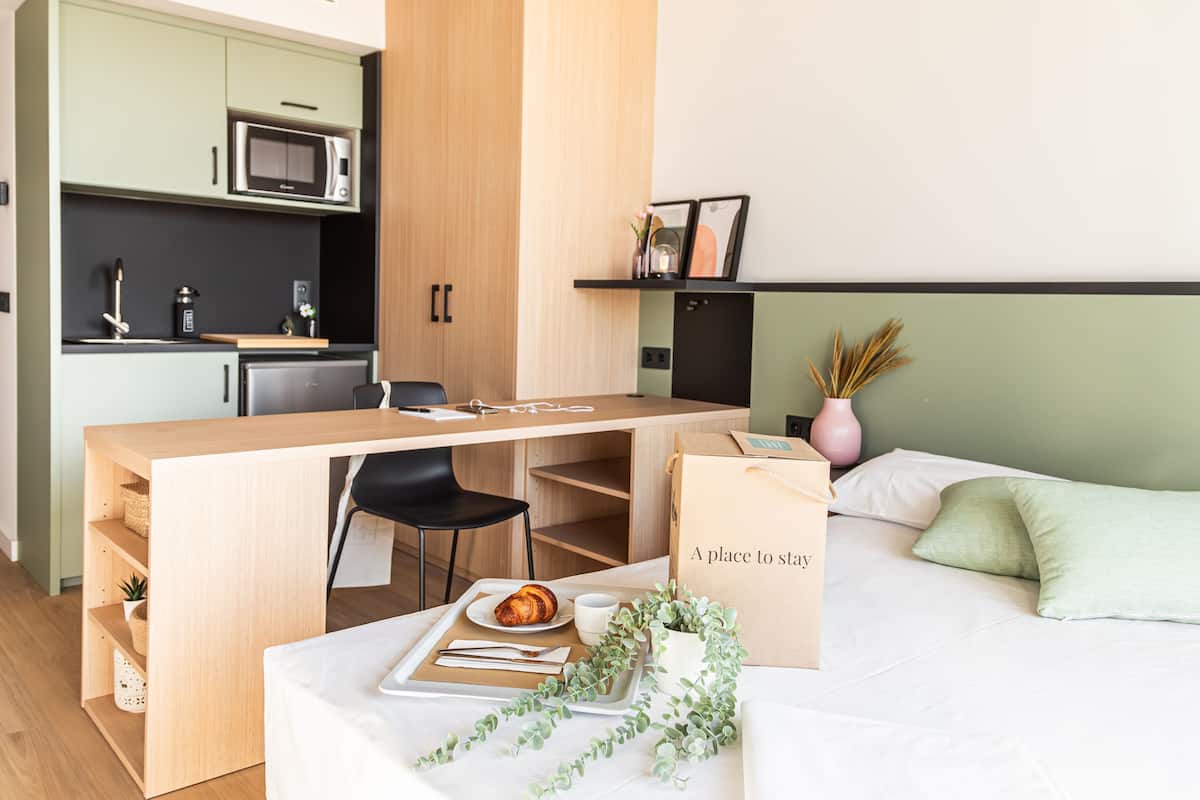A compact hotel room with a kitchenette, a bed, a desk, and a dining area.