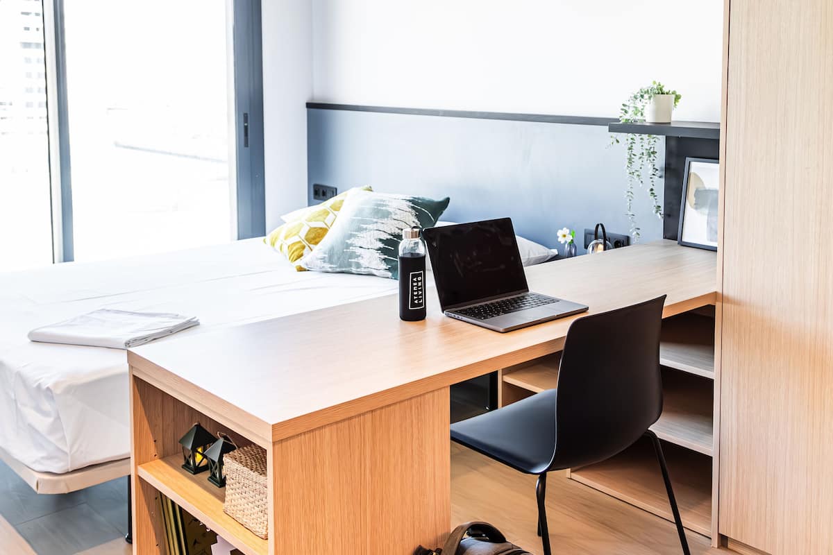 A modern bedroom with a wooden desk, a laptop, a water bottle, a bed, and a window with a city view.