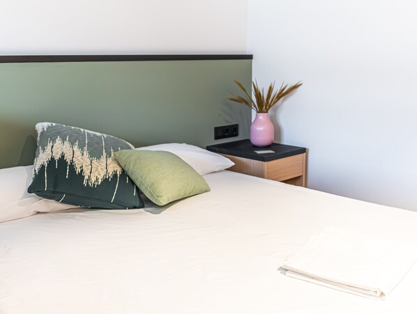A neatly made bed with two pillows, a bedside table with a vase, and a wall socket. - Atenea Living (Mataro)