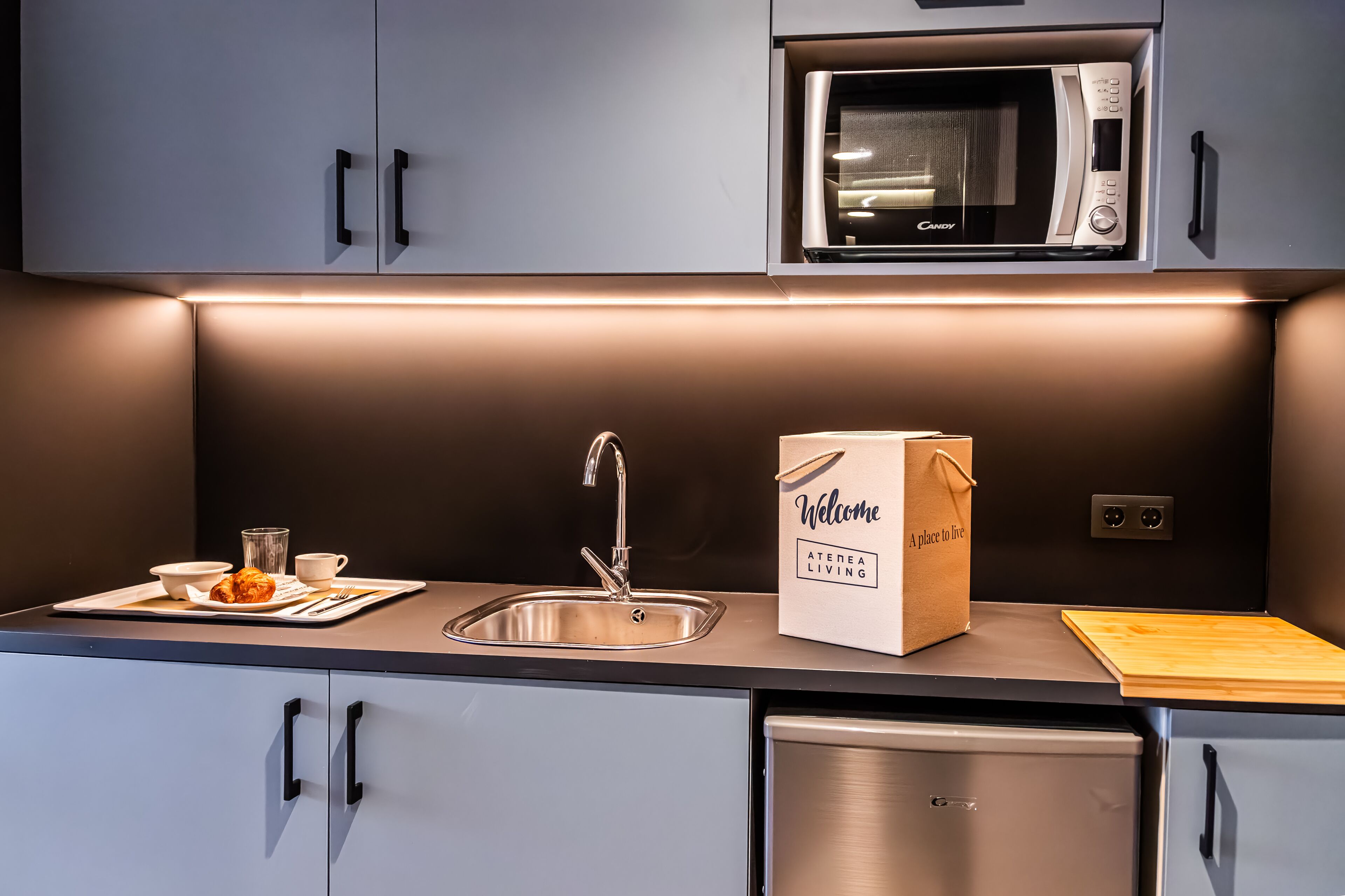 Een moderne keuken met grijze kastjes, een RVS-gootsteen, een magnetron en een houten snijplank.