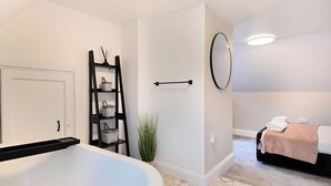 A modern bathroom with a freestanding bathtub, a black ladder shelf with baskets, a round mirror, and a bed with a beige blanket in the adjacent room.