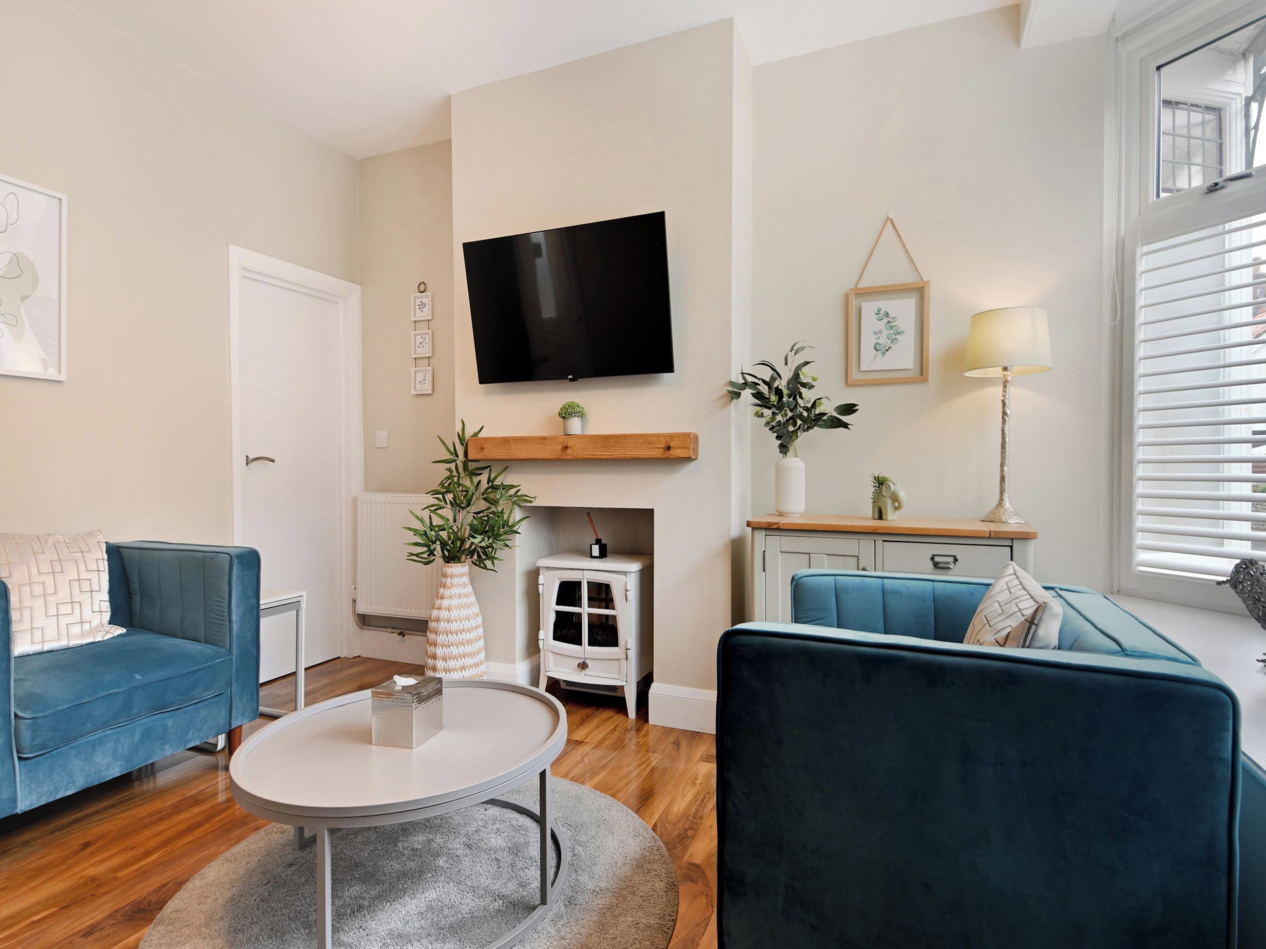 A modern living room with two blue sofas, a round coffee table, a flat-screen TV mounted on the wall, a fireplace, and a window with blinds.