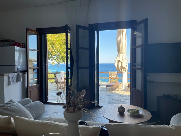A room with a white sofa, a small table with a vase, and an open door leading to an outdoor seating area with a view of the sea.