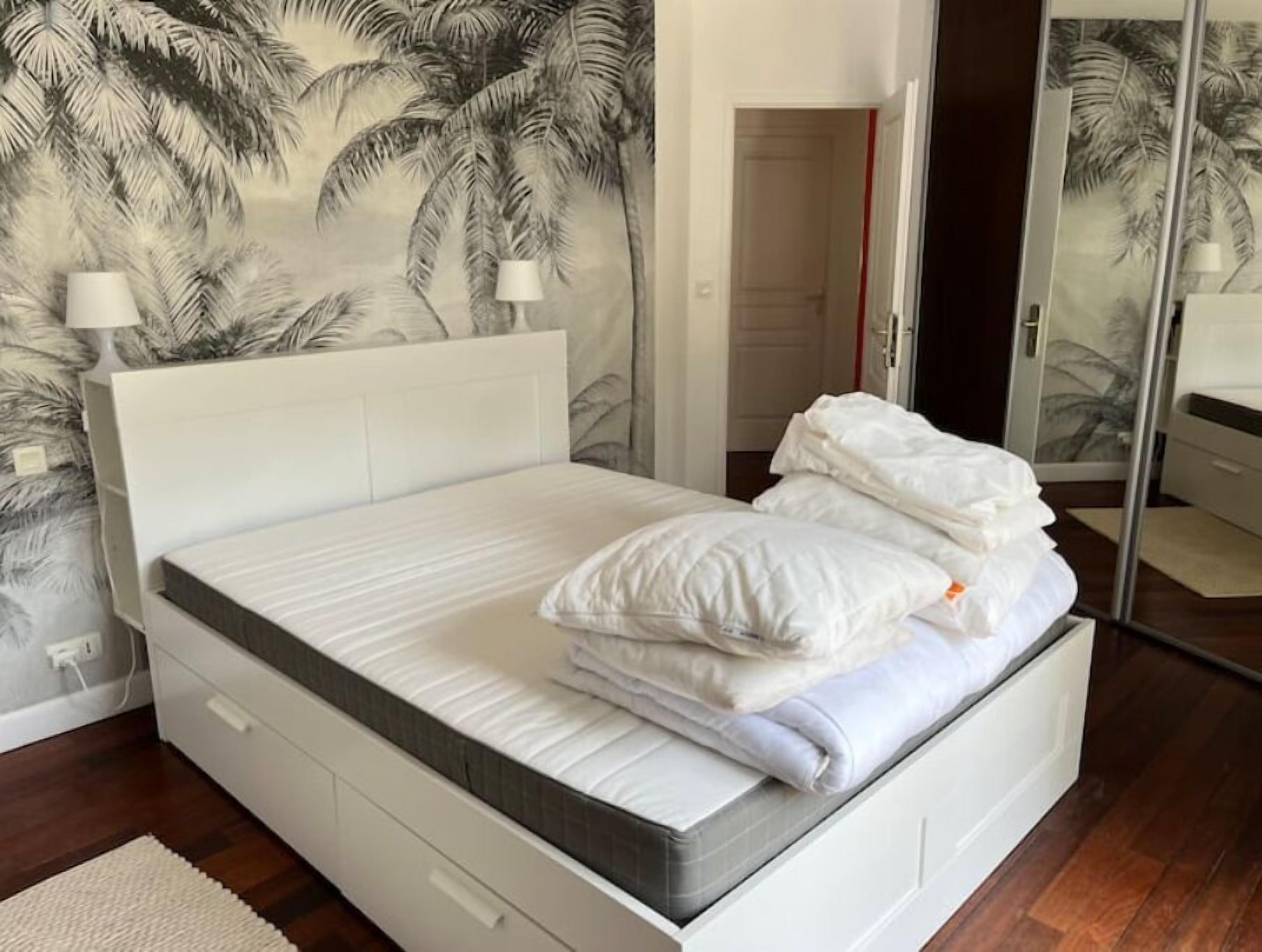 Una cama bien hecha con sábanas y almohadas blancas, un cabecero blanco y una pared con un mural de palmeras.