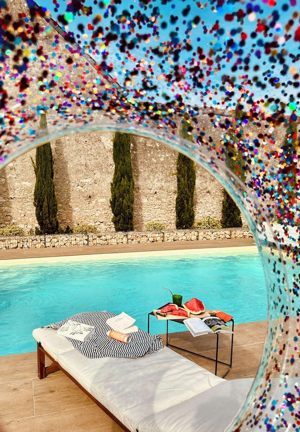 Un área de descanso junto a la piscina, con un banco, una mesa con aperitivos y vistas a un muro de piedra con árboles.