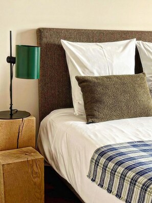 Una cama bien tendida con ropa de cama blanca y un plaid a rayas, una mesita de noche de madera con una lámpara y un cabecero texturizado.