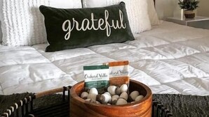 A bed with white bedding, a dark green pillow with the word "grateful," and a wooden tray with chocolate-covered nuts and two chocolate bars. - Lakeside Boho Bungalow on Table Rock Lake (Kimberling City)