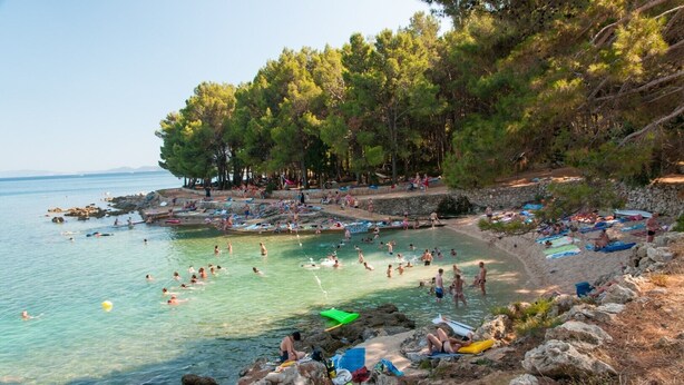 Una playa con agua cristalina, gente nadando y tomando el sol, y una costa rocosa con árboles.
