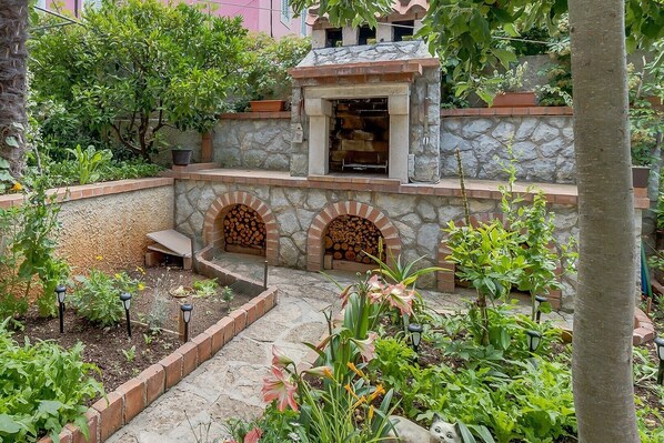 A stone patio with a fire pit, surrounded by a garden with various plants and flowers.