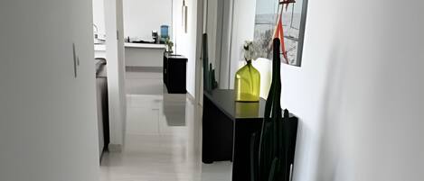 Un pasillo moderno con una zona de cocina al fondo, que incluye una mesa de comedor y sillas, y una planta en maceta.