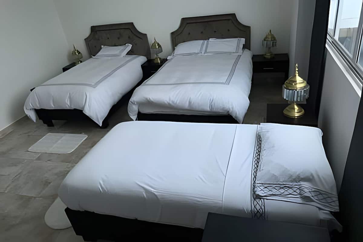 A hotel room with two single beds and one double bed, each with white bedding. There are bedside tables with lamps, a window with a view, and a small rug on the floor.