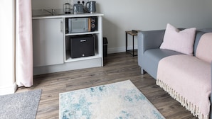 Un salon moderne avec un canapé gris, un téléviseur à écran plat fixé au mur, un coin cuisine équipé d’appareils électroménagers et un plancher en bois.