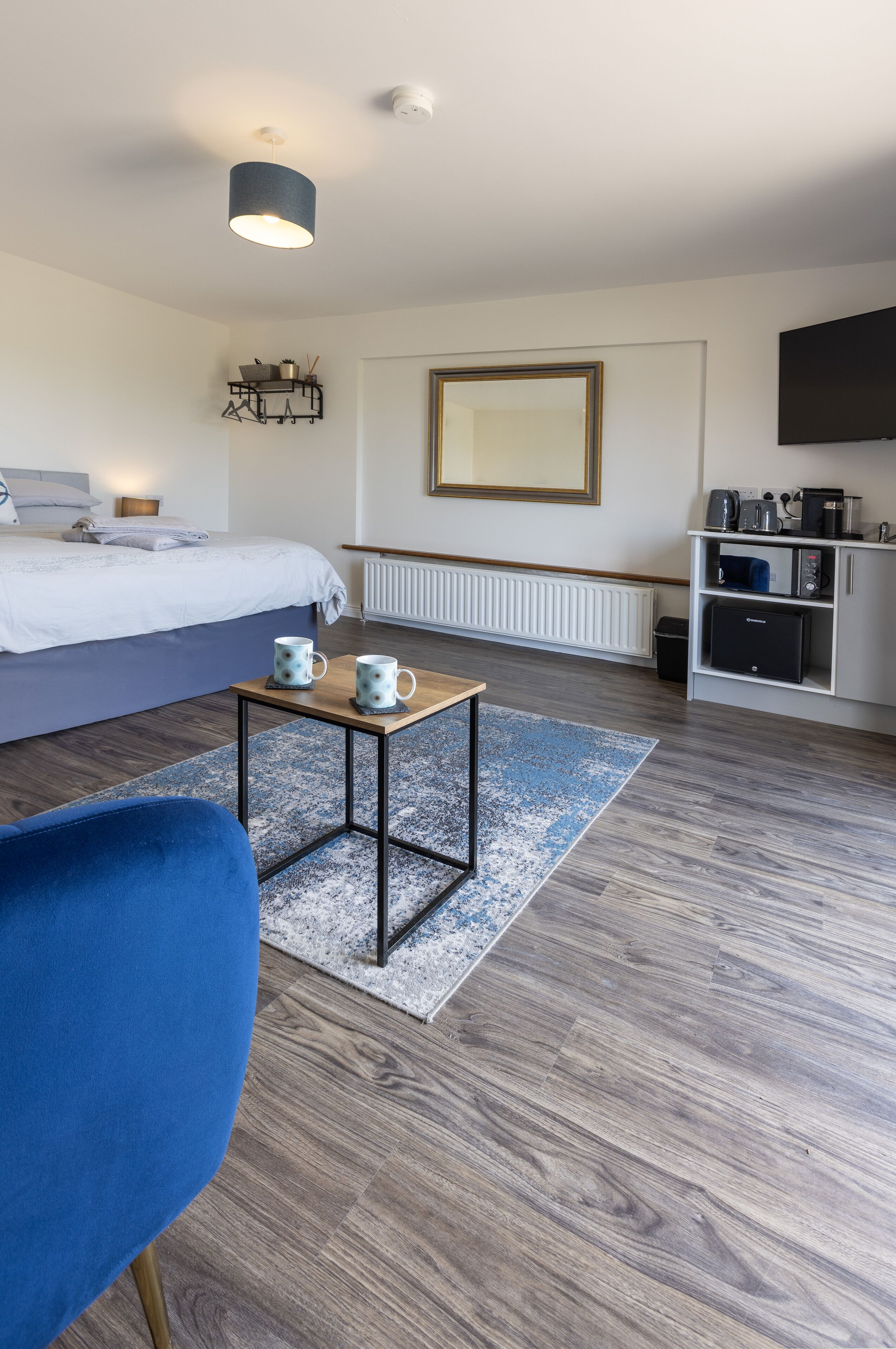 Una habitación de hotel moderna con una cama, una mesita con dos tazas, una silla azul, suelo de madera, un radiador y una kitchenette.