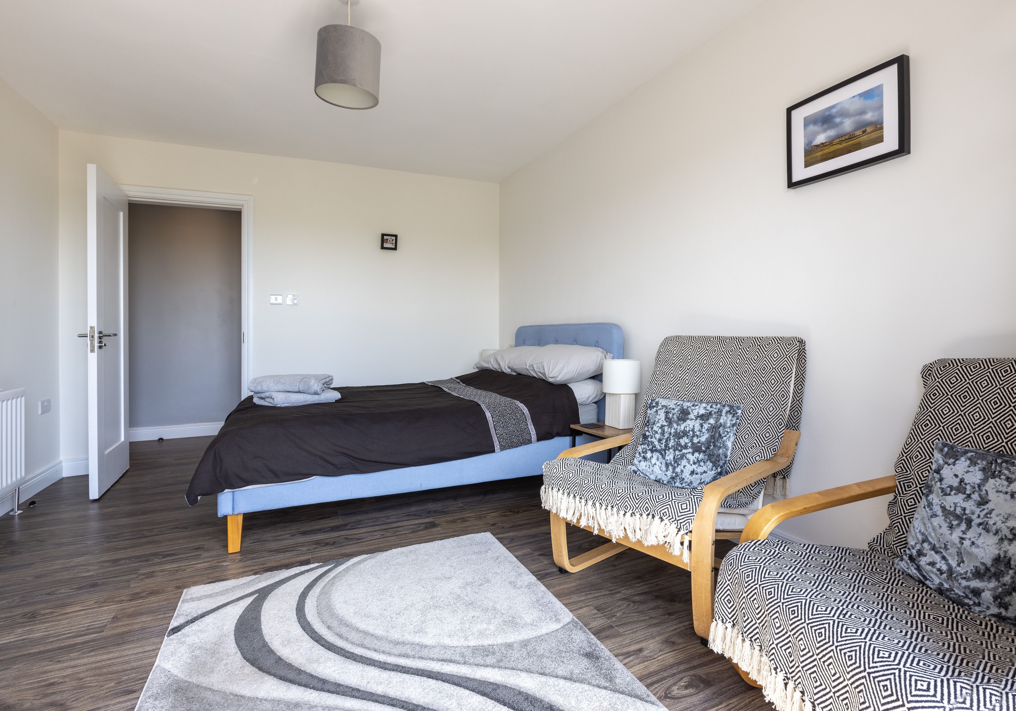 Un dormitorio con cama, dos sillas estampadas, un tapete, un cuadro en la pared y una puerta que da a otra habitación.