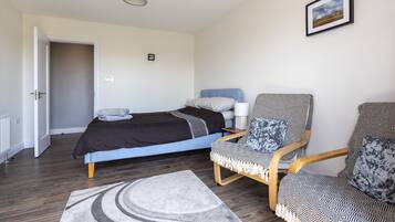 Un dormitorio con cama, dos sillas estampadas, un tapete, un cuadro en la pared y una puerta que da a otra habitación.