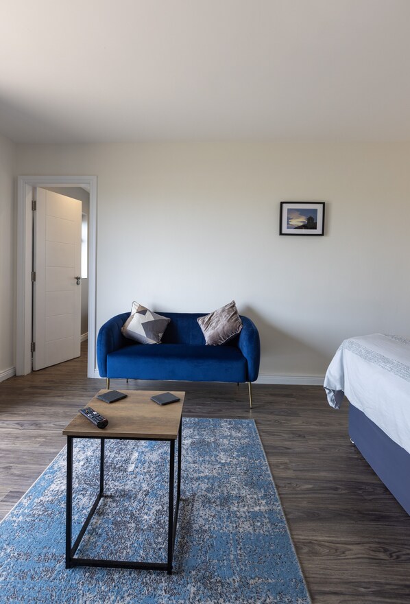 Una habitación con un sofá azul, una mesita con dos mandos a distancia, una cama, un cuadro enmarcado en la pared y una puerta que da a otra habitación.