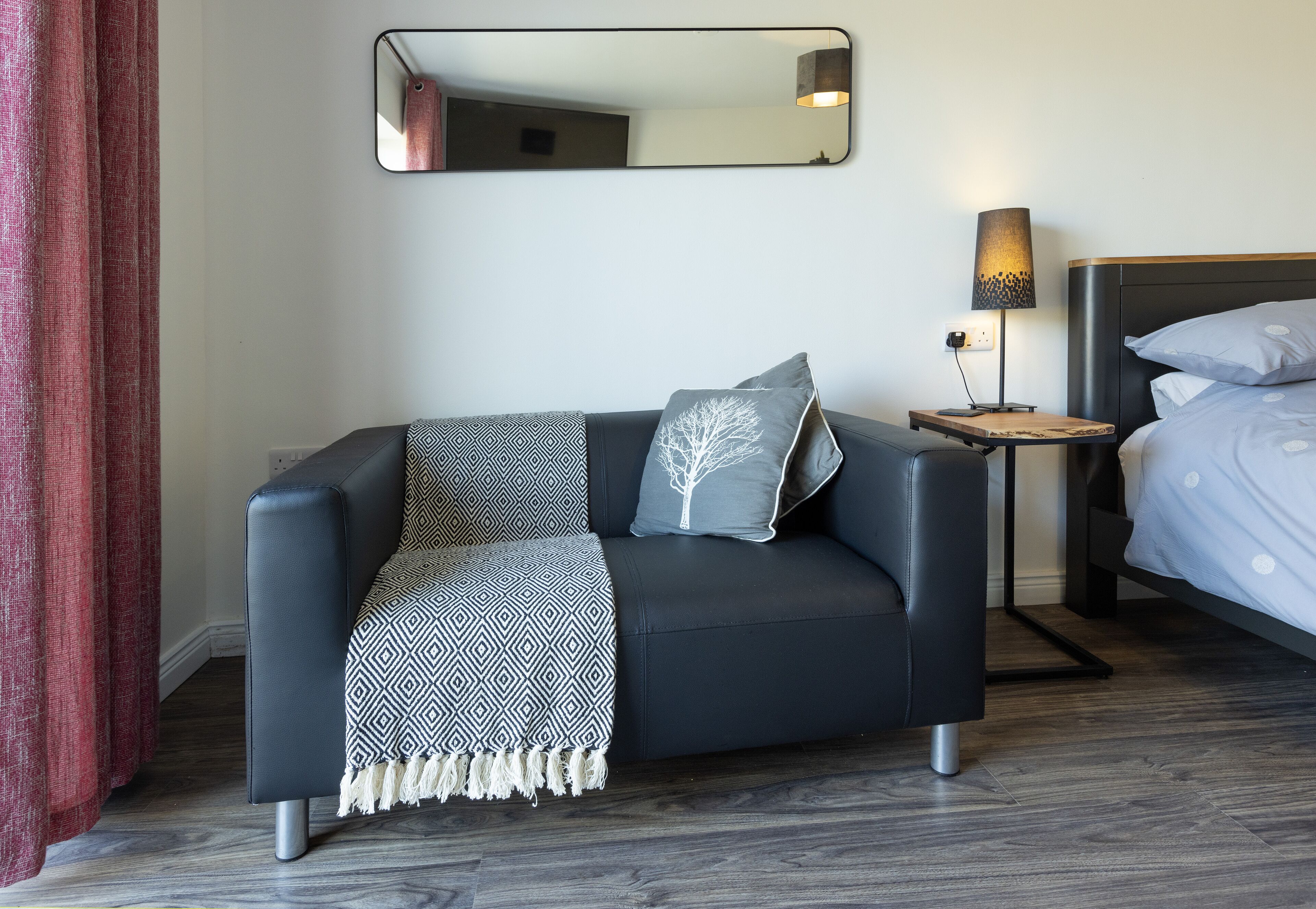 Een moderne hotelkamer met een donker leren bank, een gebreid plaid, een bijzettafeltje met een lamp, en een bed met witte beddengoed.