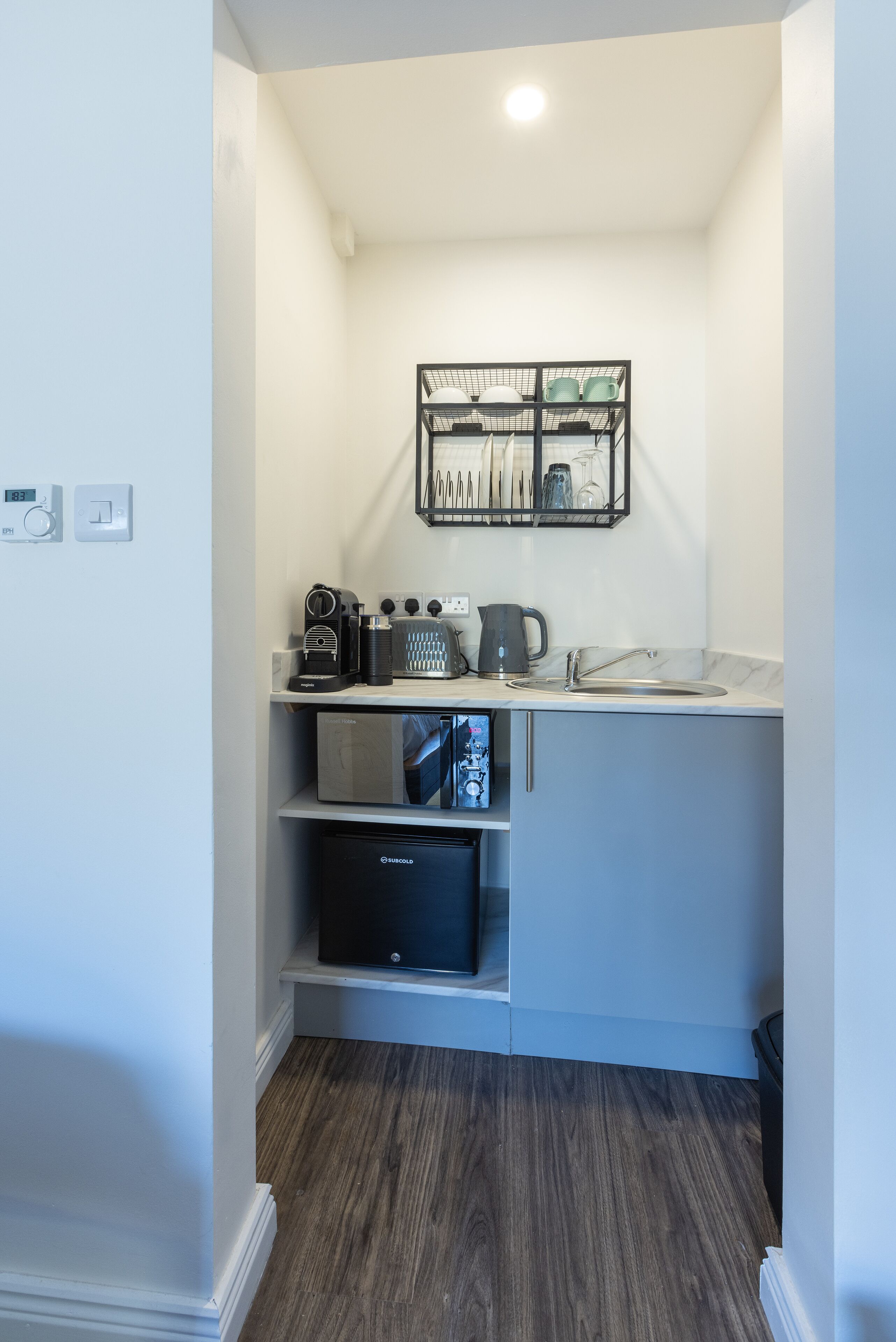A compact kitchen with a built-in coffee machine, kettle, and storage shelves.