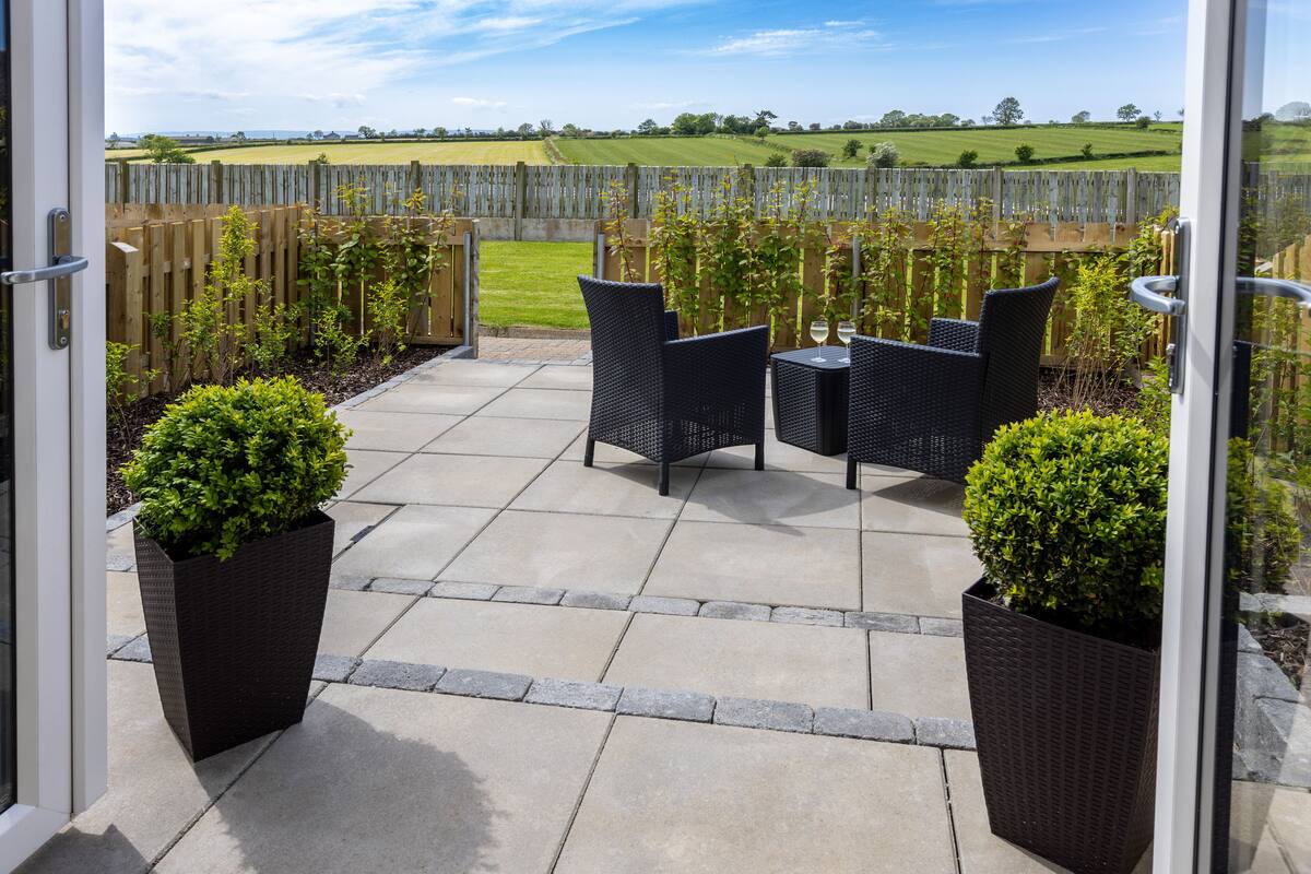 Une terrasse extérieure avec des meubles en osier et des plantes en pot, une clôture en bois et une vue sur un champ verdoyant.