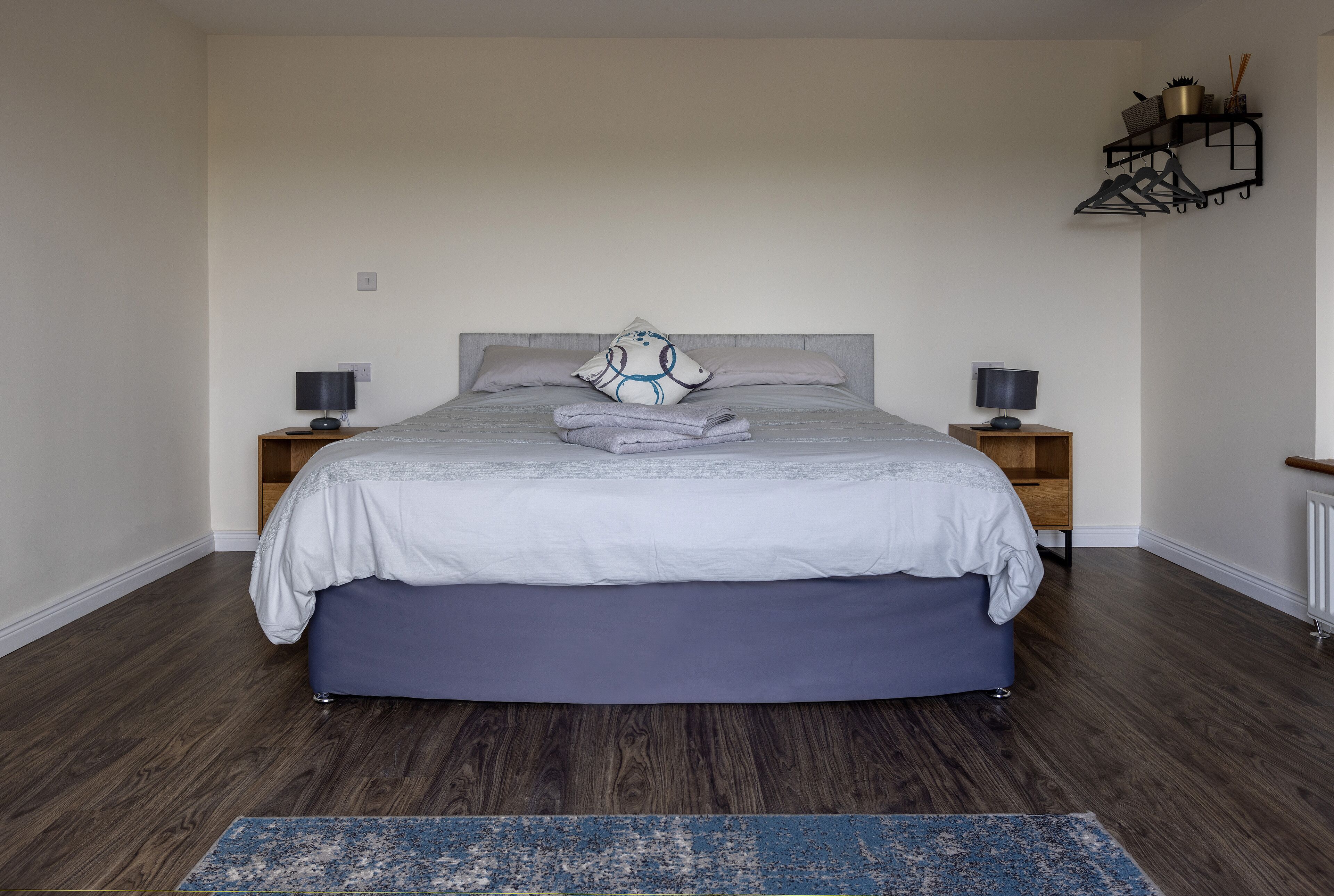 Un dormitorio con una cama, dos mesitas de noche, una mesilla auxiliar, una alfombra azul y un estante con objetos.