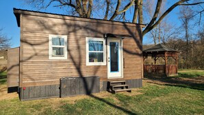 A small wooden cabin with a blue door and two windows, a wooden deck, and a gazebo in the background. - Tiny Home at Camp Mahanaim - New Paris, OH   -  Private Lake  (New Paris)