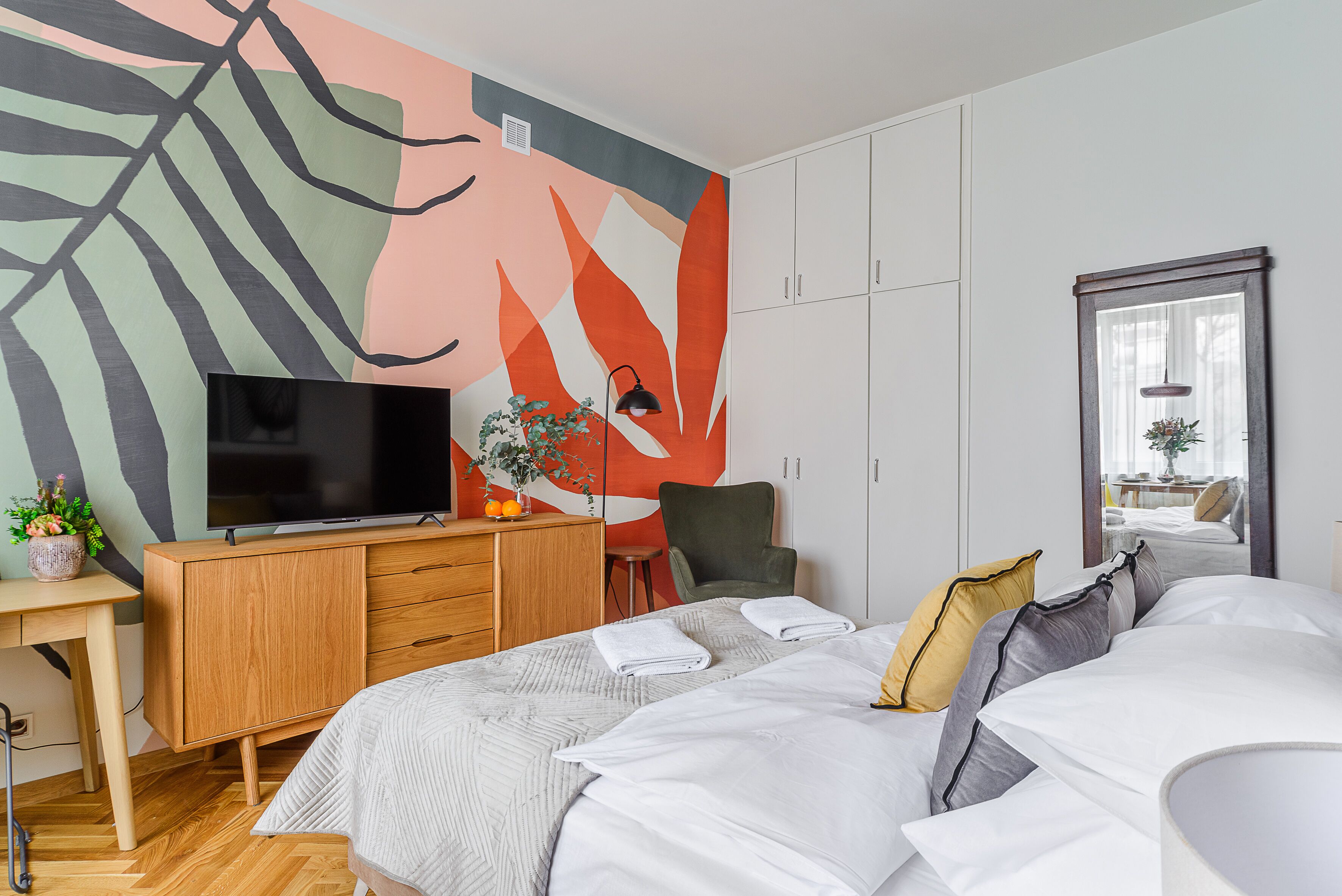 A modern bedroom with a large bed, a desk, a TV, and a vibrant abstract wall mural.