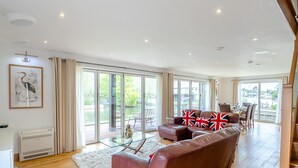 A spacious living room with a large sofa, a glass coffee table, and a view of the outdoors through large sliding doors. - This stunning waterside home offers an amazing space for families and friends. (Wroxham)