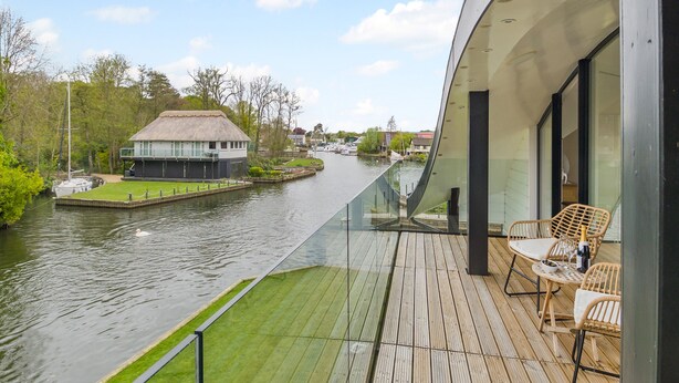 A modern building with a glass balcony overlooking a river, a thatched-roof structure, and a wooden deck with outdoor furniture. - This stunning waterside home offers an amazing space for families and friends. (Wroxham)