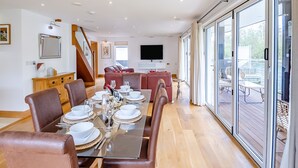 A spacious dining area with a long table set for a meal, a comfortable seating area with sofas, a staircase, and a large sliding glass door leading to an outdoor deck. - This stunning waterside home offers an amazing space for families and friends. (Wroxham)