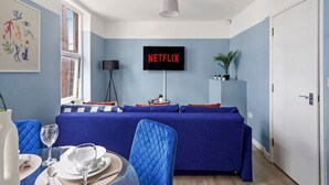 A modern living room with a blue sofa, a dining table with chairs, a flat-screen TV displaying "NETFLIX", and a window providing natural light.