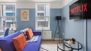 A modern living room with a blue sofa, orange cushions, a glass coffee table, and a large TV screen displaying "NETFLIX".
