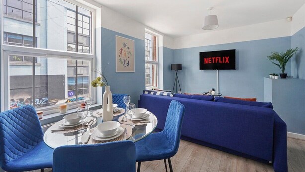 A modern living room with a blue sofa, a dining table set with plates and glasses, and a large window with a view of buildings.