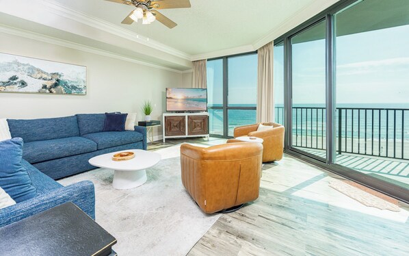 A modern living room with a blue sofa, a white coffee table, and a leather armchair. A flat-screen TV is mounted on a wooden cabinet, and the room has large windows offering a view of the ocean.
